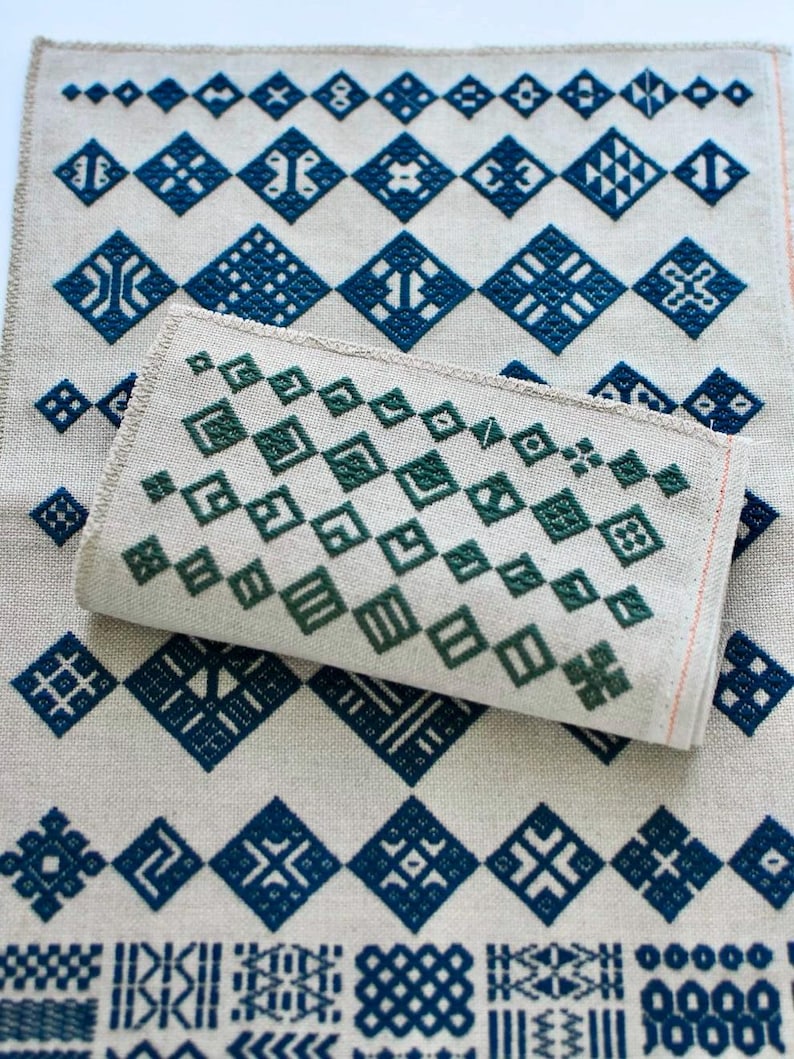 May include: A rectangular piece of light beige fabric featuring a repeating pattern of dark blue geometric diamond shapes and intricate symbols. A smaller, folded piece of the same fabric with dark green diamond patterns rests on top.
