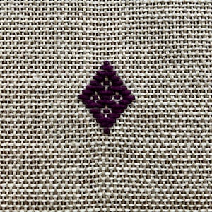 May include: A piece of light beige, coarse-woven fabric featuring a single, small, dark purple diamond-shaped embroidery pattern centered in the frame. The embroidery consists of thick, diagonal stitches that form a geometric, open-work design.