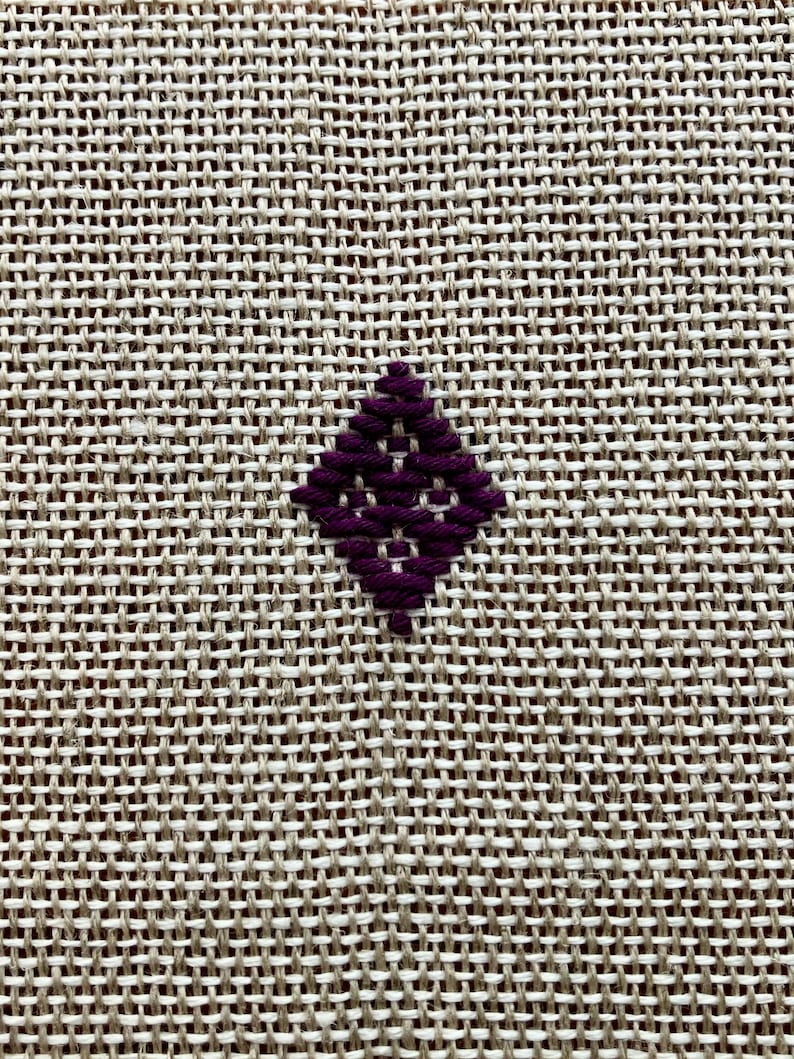 May include: A piece of light beige, coarse-woven fabric featuring a single, small, dark purple diamond-shaped embroidery pattern centered in the frame. The embroidery consists of thick, diagonal stitches that form a geometric, open-work design.