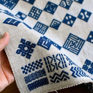 May include: A beige fabric runner featuring a grid of dark blue embroidered geometric patterns. Each square contains a unique design, including zigzags, lines, and cross-hatch motifs. The edges of the fabric are frayed, showing a woven texture.