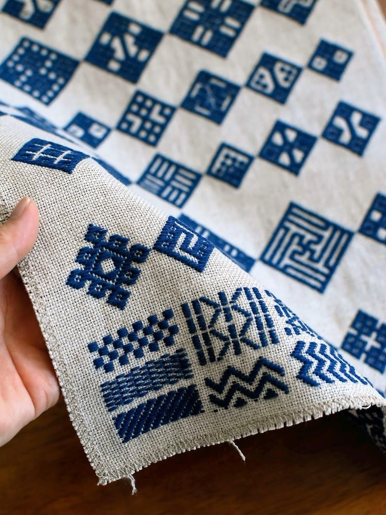 May include: A beige fabric runner featuring a grid of dark blue embroidered geometric patterns. Each square contains a unique design, including zigzags, lines, and cross-hatch motifs. The edges of the fabric are frayed, showing a woven texture.