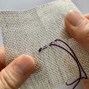 May include: A square piece of light beige, open-weave fabric with a grid-like texture. A single strand of deep purple embroidery thread is being stitched through the fabric, creating a small, horizontal line of stitches on the surface.