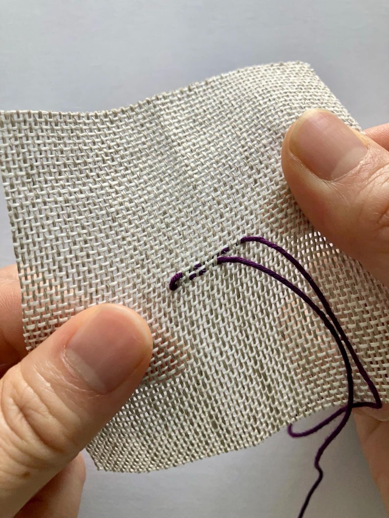 May include: A square piece of light beige, open-weave fabric with a grid-like texture. A single strand of deep purple embroidery thread is being stitched through the fabric, creating a small, horizontal line of stitches on the surface.
