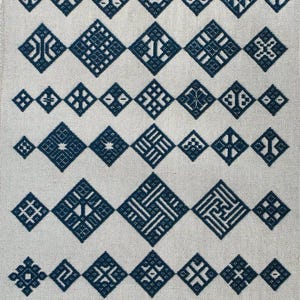 May include: A piece of light beige fabric featuring a collection of dark blue cross-stitch embroidery patterns. The design consists of multiple rows of geometric diamond shapes, each containing unique, intricate, and symmetrical line motifs.