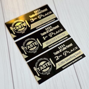 May include: Black and gold award certificates from the 2024 Taste of Lake Forest event. The certificates display the event logo, place rankings, and restaurant names. The certificates are arranged on a light wood surface.
