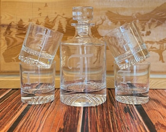 May include: A set of four clear glass decanter and glasses with a single initial etched on each piece. The decanter has a stopper and the glasses have a textured rim.
