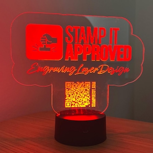 May include: Red acrylic sign with a white cloud shape, featuring the text "STAMP IT APPROVED" and a thumbs up icon. The sign also includes the text "Engraving Laser Design" and a QR code with the website "STAMPITESTY.COM". The sign is illuminated from below by a red LED base.
