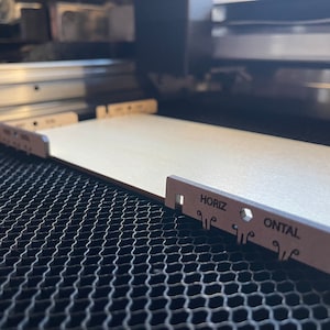 May include: A laser cutting machine in operation, with a light-coloured wooden plank in place. Metal guides hold the wood, and the text "HORIZ ONTAL" is visible on one of them. The image is taken from a close-up perspective.