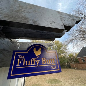 以下が含まれることがあります： 「The Fluffy Butt Hut」と金色の文字で書かれた青い木製の看板。テキストの上に金の鶏のシルエットがあります。看板は木製の構造物にぶら下がっています。