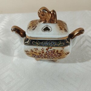 May include: A small, white porcelain incense burner with a gold-painted lid and a gold-painted animal figure on top. The burner has a square base and is decorated with floral patterns in gold, red, and blue.