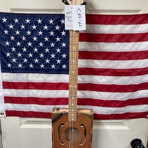 May include: A handmade cigar box guitar with a wooden neck and fretboard. The guitar's body is a brown cigar box with decorative cutouts. The headstock has tuning pegs. The guitar is displayed in front of an American flag.