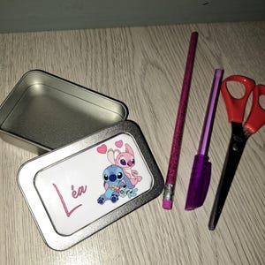 Peut inclure: Une boîte en métal argentée avec un autocollant blanc représentant un couple de personnages de dessin animé bleu et rose avec des cœurs. Le nom "L'ea" est écrit en cursive rose sur l'autocollant.