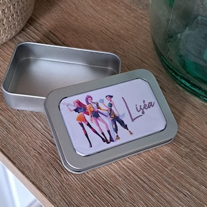 Peut inclure: Petite boîte rectangulaire argentée ouverte avec un couvercle. Le couvercle présente une illustration colorée de trois personnages et le nom "Liséa" en écriture violette. La boîte est posée sur une surface en bois.
