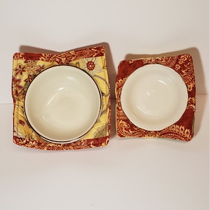 May include: Two quilted bowl cozies in red and yellow floral fabric. The cozies are square and have a brown trim around the opening.
