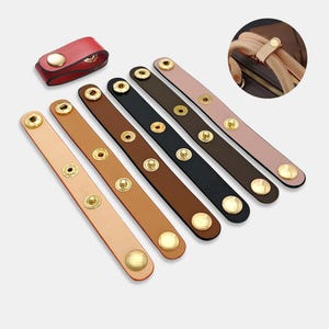 May include: A collection of leather straps in various colors, including tan, brown, black, and pink, each with gold-colored snap closures. A small red leather key holder with a gold snap is also present. These straps are designed for bags and accessories.