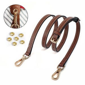 May include: A brown leather purse strap with white stitching and gold-colored metal hardware. The strap has a buckle for adjustment and two swivel clasps. Also included are six gold-colored eyelets. A close-up shows the strap attached to a brown purse.