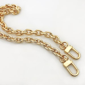 Peut inclure: Une chaîne dorée avec des maillons ovales et deux fermoirs mousquetons. La chaîne est en métal et est conçue pour être utilisée comme sangle pour un sac à main. Les fermoirs sont attachés à la chaîne.