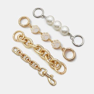 Peut inclure: Quatre lanières décoratives pour sacs à main en or, argent et blanc. L'une présente de grosses maillons dorés, une autre une série de perles blanches et une troisième des breloques dorées en forme de pattes. La quatrième lanière a de petites maillons dorés.