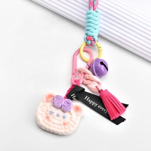 Puede incluir: Llavero de oso de crochet rosa con lazos morados, un borla rosa, una campana morada y una cinta negra con el texto "Happy every".