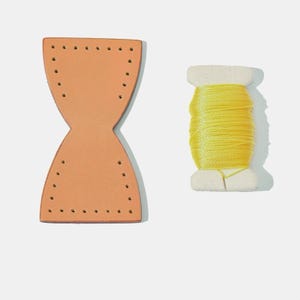 May include: A light brown leather piece with pre-punched holes for stitching, next to a spool of bright yellow thread. The leather piece has an hourglass shape. The thread is wound around a white spool.