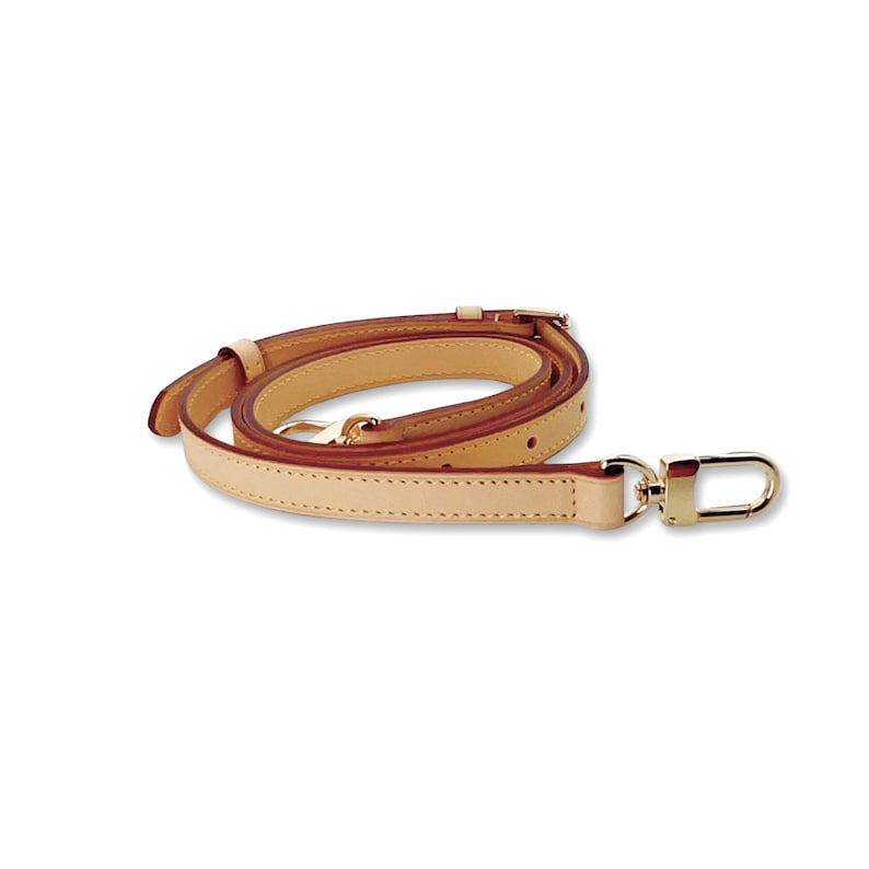 May include: A tan leather bag strap with gold-colored hardware. The strap is made of a light tan leather with a darker brown edge. The strap has a gold-colored clasp on one end and a gold-colored buckle on the other end.