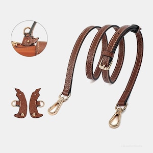 May include: A brown leather handbag strap with gold-colored metal clasps and a buckle. The strap is shown coiled, with white stitching along the edges. Two additional leather pieces with metal rings are also shown.