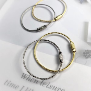 May include: Two keychains, one silver and one gold, are intertwined on a white surface. The keychains are made of a flexible, coiled wire with a screw-on clasp. The gold keychain has a textured clasp, while the silver one has a smooth clasp.
