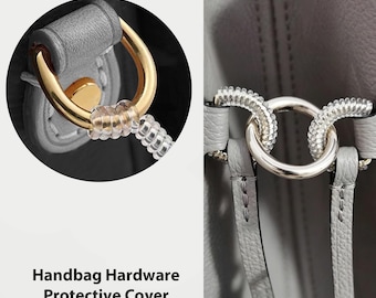 May include: Close-up of handbag hardware, showcasing a gold-toned ring and a silver-toned ring. The gold ring is connected to a clear, coiled protective cover. The text "Handbag Hardware Protective Cover" is at the bottom.