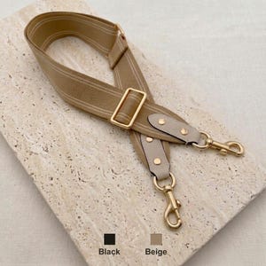 Peut inclure: Une bandoulière de sac en tissu beige avec de fines rayures blanches, un curseur réglable en métal doré et deux mousquetons pivotants dorés fixés sur des pattes en similicuir beige clair avec des rivets dorés. Le texte Black and Beige est visible.