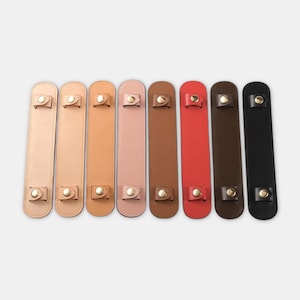 Puede incluir: Una colección de ocho almohadillas de cuero para correas de hombro en varios colores, incluyendo beige, marrón claro, rosa, marrón, rojo y negro. Cada almohadilla tiene una parte superior e inferior redondeadas con herrajes metálicos dorados.