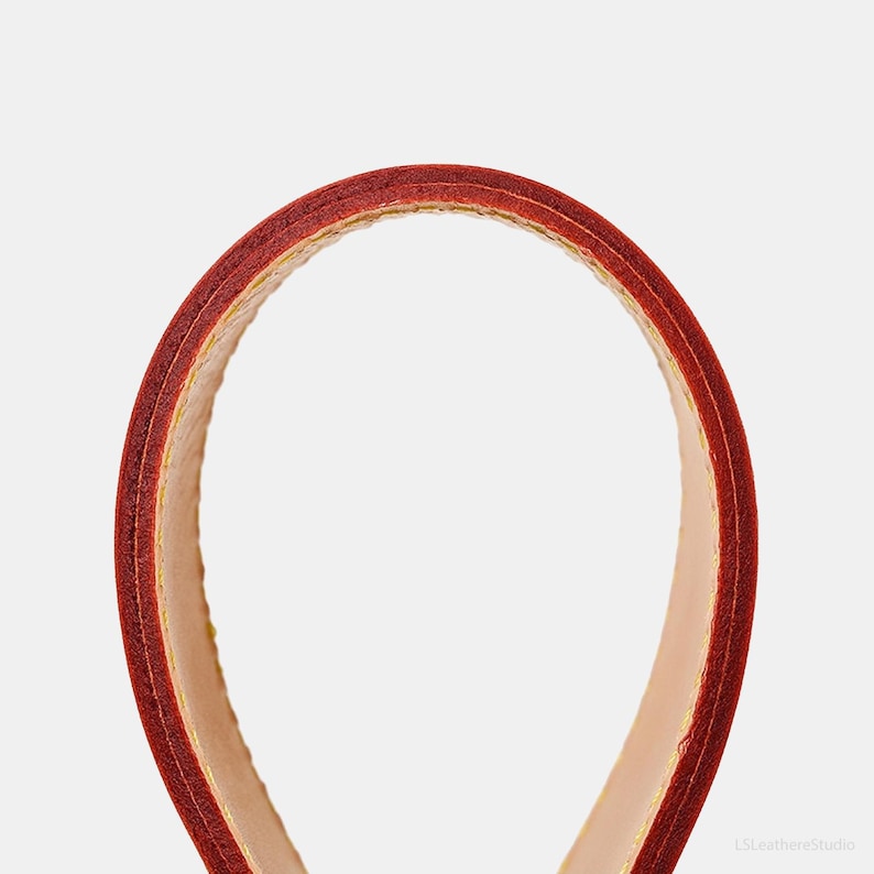 May include: Close-up of a curved, red leather handle with a light beige interior and yellow stitching. The handle is likely from a bag or purse, showcasing the craftsmanship and materials used in its construction.