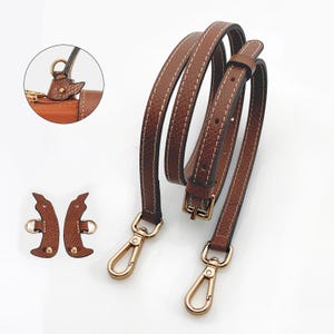 Könnte beinhalten: Brauner Leder-Handtaschenriemen mit weißen Nähten und goldfarbener Metallbeschlägen. Enthält zwei zusätzliche Lederstücke mit Metallringen und eine Nahaufnahme des Riemenanschlusses.