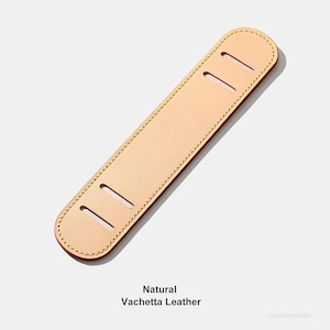 May include: A natural Vachetta leather shoulder pad with three rectangular cutouts. The edges are stitched with a contrasting thread, and the leather has a smooth, light tan appearance. The text "Natural Vachetta Leather" is at the bottom.