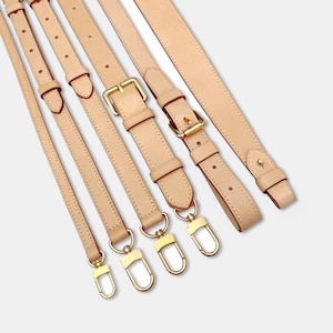 Puede incluir: Varias correas de bolso de cuero beige con herrajes metálicos dorados están dispuestas en diagonal. Las correas varían en longitud e incluyen hebillas ajustables y cierres giratorios. El cuero parece liso y ligeramente texturizado.