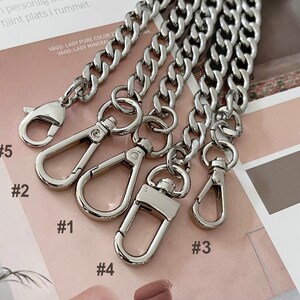 Puede incluir: Primer plano de varias correas de cadena de color plateado con varios estilos de cierre. Las cadenas son gruesas y tienen un dise&ntilde;o de eslabones de bordillo. Los cierres incluyen estilos de mosquet&oacute;n y gancho giratorio. El fondo es una superficie impresa rosa y marr&oacute;n.