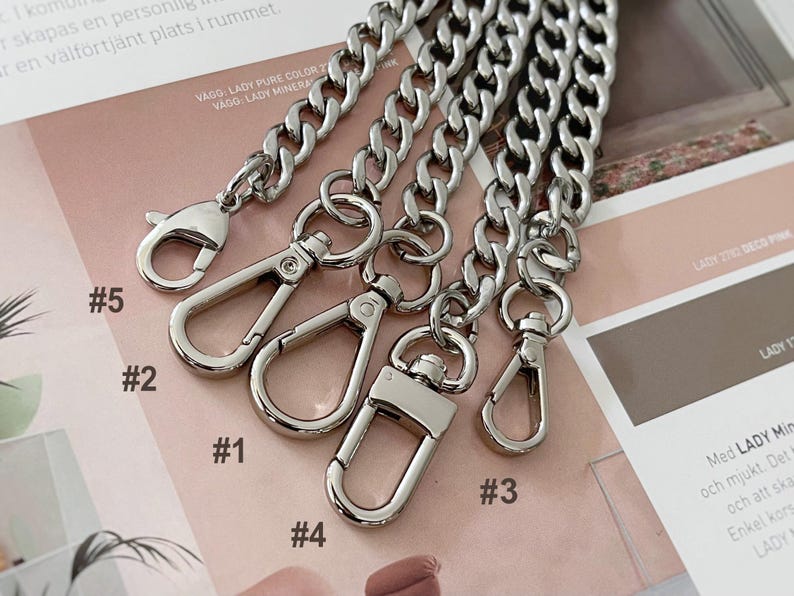 Puede incluir: Primer plano de varias correas de cadena de color plateado con varios estilos de cierre. Las cadenas son gruesas y tienen un dise&ntilde;o de eslabones de bordillo. Los cierres incluyen estilos de mosquet&oacute;n y gancho giratorio. El fondo es una superficie impresa rosa y marr&oacute;n.