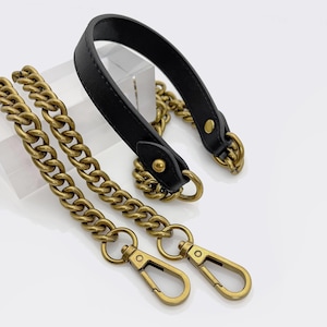 May include: A black leather handbag handle with gold-coloured metal chain straps and clasps. The chain links are thick and the clasps are a simple, functional design. The handle is attached to the chain with gold-coloured metal rings.