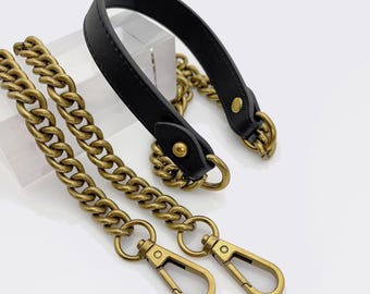 May include: A black leather handbag handle with gold-colored metal chain straps and clasps. The chain links are thick and the clasps are a simple, functional design. The handle is attached to the chain with gold-colored metal rings.