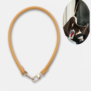 May include: A tan leather handbag strap with gold-coloured metal hardware. The strap is a rounded shape and has a clasp at each end for easy attachment to a purse or bag. The strap is shown against a white background.