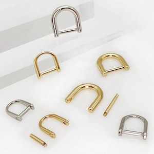 Peut inclure: Divers assortiments d'anneaux en D métalliques de tons or et argent. Les pièces de quincaillerie sont disposées sur une surface blanche, mettant en valeur leur forme incurvée et leur design en barre. Idéal pour la maroquinerie et autres projets d'artisanat.