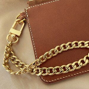 Puede incluir: Una cartera de cuero marr&oacute;n con una correa y cierre de cadena dorada. La cartera tiene costuras blancas alrededor de los bordes. La cadena dorada es de estilo cadena de bordillo.