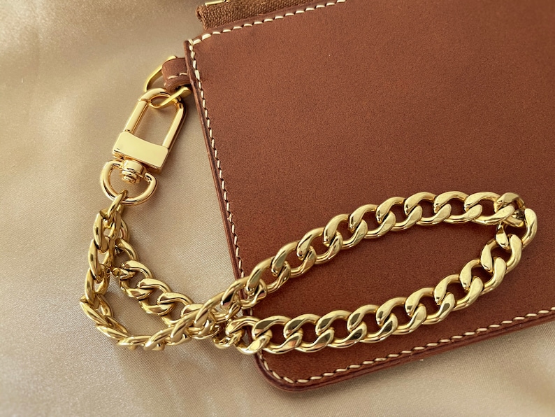 Puede incluir: Una cartera de cuero marr&oacute;n con una correa y cierre de cadena dorada. La cartera tiene costuras blancas alrededor de los bordes. La cadena dorada es de estilo cadena de bordillo.