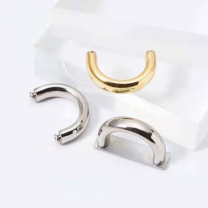 May include: Three metal cabinet pulls in a U-shape. One is gold, and two are silver. The silver pulls have a cylindrical shape with screw attachments on each end. The gold pull is a smooth, curved design. All three are on a white background.