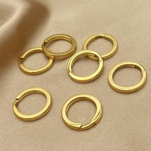 May include: A collection of gold-colored key rings. The rings are circular with a hinged opening, and are arranged on a neutral-toned fabric surface. The key rings are made of a shiny, metallic material.