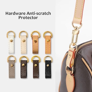 Pode incluir: Imagem de protetores anti-riscos para hardware em várias cores, incluindo branco, bege, castanho claro e preto. Cada protetor apresenta um anel de metal dourado e uma tira de couro com um rebite dourado. Os protetores são projetados para serem fixados à alça de uma bolsa.