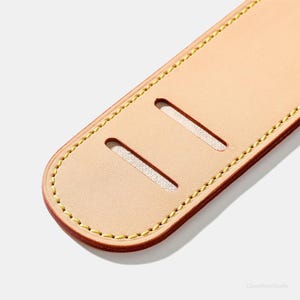 May include: Close-up of a light tan leather strap with two rectangular cutouts. The edges are stitched with yellow thread, and the border is a darker brown. The strap is likely for a bag or other accessory.