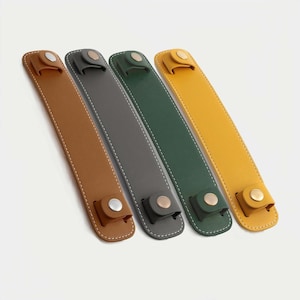 May include: Four leather straps in shades of brown, grey, green, and yellow. Each strap has a button closure and white stitching along the edges. The straps are arranged diagonally against a white background.