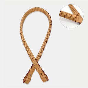 May include: A tan leather bag strap featuring a braided design throughout the main length. The strap ends in two flat leather tabs, each secured with a single metallic stud, allowing for attachment to a bag. A circular inset shows the braid detail.