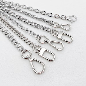 Puede incluir: Una colección de cinco cadenas de metal en tono plateado dispuestas en abanico. Cada cadena presenta un acabado pulido y está equipada con un cierre de mosquetón en el extremo, diseñado para una fijación segura a diversos accesorios.