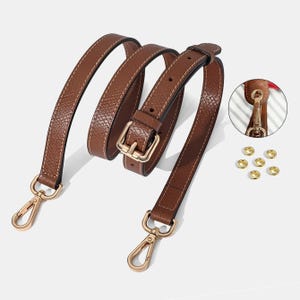 May include: A brown leather bag strap with gold-colored metal hardware, including clasps and a buckle. The strap is stitched with a lighter thread and has a textured surface. Additional gold rivets are also visible.